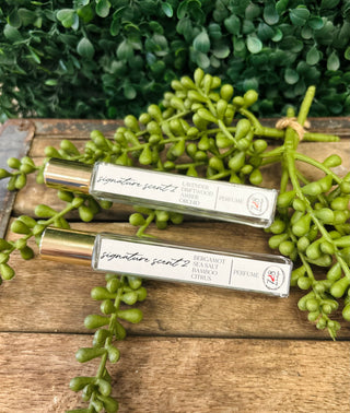 Two Vincent 728 signature perfume rollers with gold caps displayed on wooden surface with green foliage.