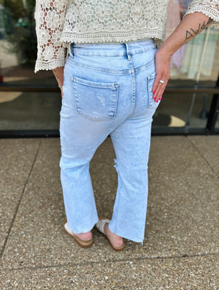 Woman wearing light blue distressed cropped straight-leg jeans with white lace top, back view showing fit and styling.