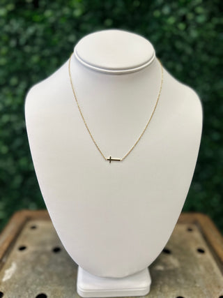Delicate gold chain necklace with small cross pendant displayed on white jewelry bust outdoors.