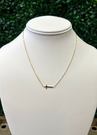 Gold cross pendant necklace displayed on white jewelry bust with green foliage background.