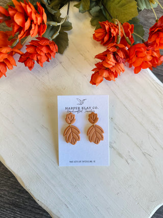 Handcrafted clay leaf dangle earrings displayed on a branded card with fall floral backdrop.