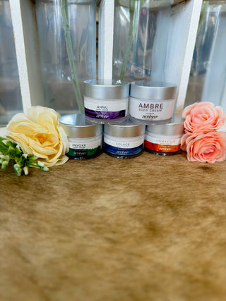Five Ambre Blends body cream jars displayed on wooden surface with decorative flowers and glass vases.