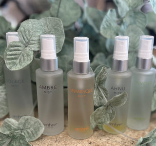 Five frosted glass spray bottles with metallic pumps arranged with decorative green leaves on textured surface.