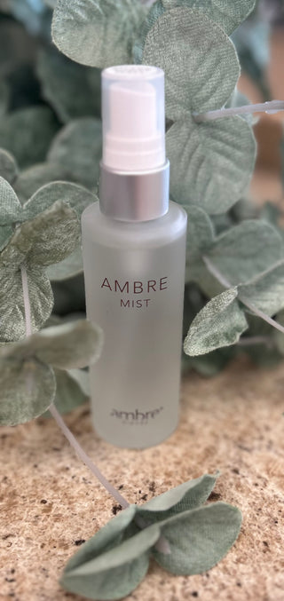 Frosted glass spray bottle of Ambre Mist surrounded by decorative sage green leaves on textured surface.