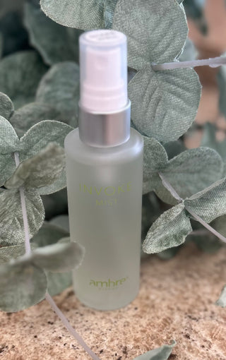 Frosted glass spray bottle with white pump surrounded by decorative sage green fabric leaves on textured surface.