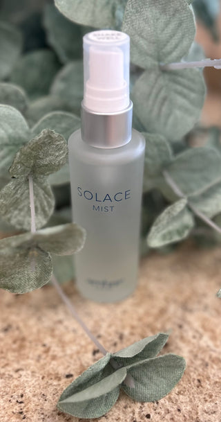 Clear spray bottle labeled "Solace Mist" positioned among green fabric leaves on textured surface.