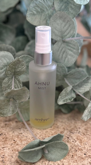Ambre Blends AHNU body mist spray bottle surrounded by decorative eucalyptus leaves on textured surface.