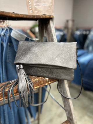 Gray leather foldover crossbody clutch with tassel detail displayed on rustic wooden stool in boutique setting.
