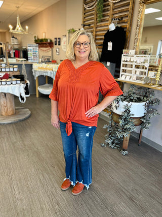 Woman wearing orange eyelet top with tie-front detail and jeans, standing in boutique store setting.