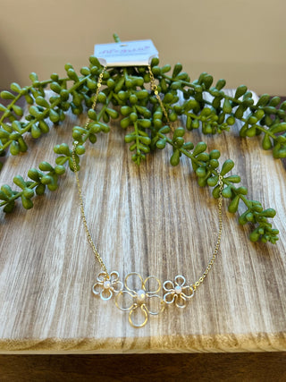 Gold floral necklace with interconnected flower motifs displayed on wooden surface with green succulent plant.