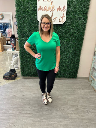 Woman wearing green v-neck tee and black leggings in boutique setting with artificial grass wall backdrop.
