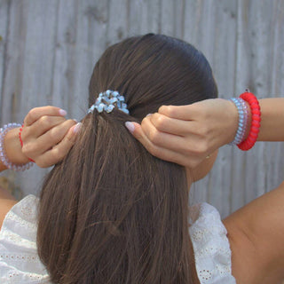 Person styling long brown hair with decorative hair tie, wearing colorful beaded bracelets against wooden fence background.
