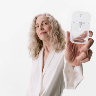 Woman in white robe holding Touchland Power Mist hydrating hand sanitizer bottle against white background.