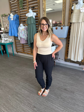 Woman wearing cream tank top and black joggers standing in boutique with clothing displays