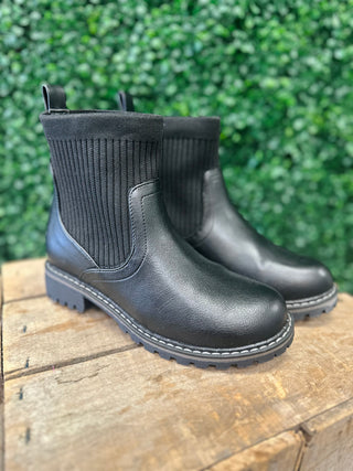 Black ankle boots with ribbed knit upper and leather lower on wooden surface with green foliage background.
