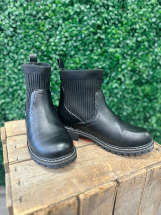 Black ankle boots with ribbed knit upper and leather lower on wooden surface with green foliage background.