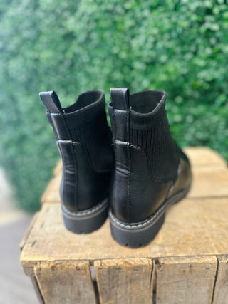 Black ankle boots with elastic side panels displayed on rustic wooden surface outdoors