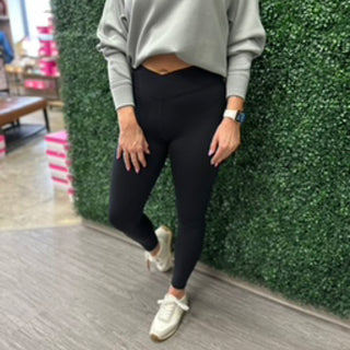 Woman wearing black high-waisted leggings with crossover waistband detail and beige sweater, standing against green backdrop.