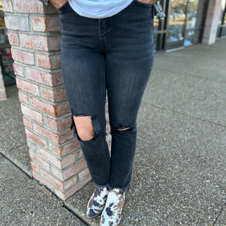 Person wearing black distressed jeans with knee rips and frayed hem, standing against brick wall.