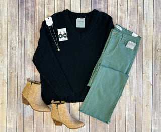 Black knit sweater outfit flatlay with green jeans, tan boots, necklace and earrings on wood background