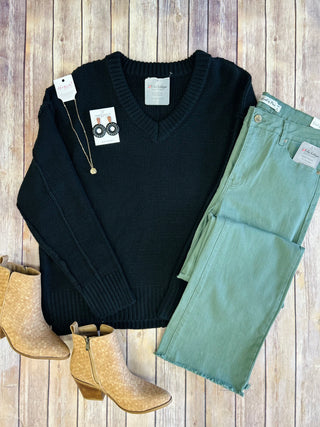 Black knit sweater with v-neck styled with green jeans, tan boots, and accessories on wooden background