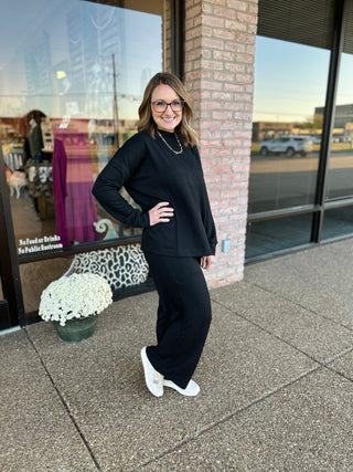 Woman wearing black ribbed mock neck sweater and matching pants set standing outside storefront