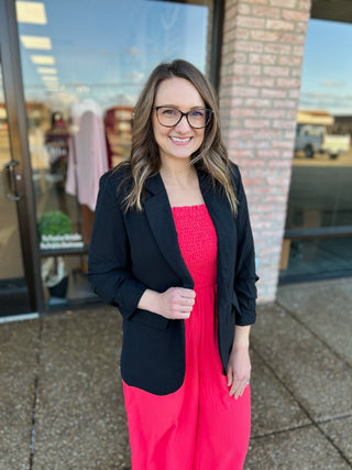 Woman wearing black ruched sleeve blazer over red dress, standing outdoors near brick building storefront.
