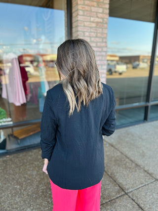 Woman wearing navy blue ruched sleeve blazer with pink pants, shown from behind in boutique setting.