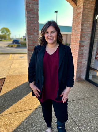 Woman wearing black ruched sleeve blazer over burgundy top, standing outdoors near brick archway.