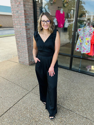 Woman wearing black sleeveless wide leg jumpsuit with v-neck, standing outside storefront with glasses and pearl necklace.