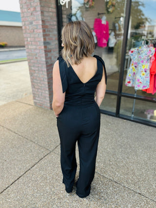 Woman wearing black wide leg jumpsuit with tie shoulder straps, shown from behind on sidewalk.