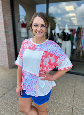 Woman wearing colorful patchwork top with floral and geometric patterns, standing outside boutique storefront.