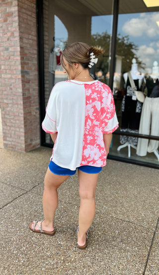 Woman wearing white and pink floral colorblock top with blue shorts, viewed from behind near storefront.