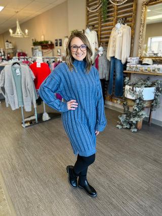 Woman wearing blue cable-knit sweater dress in boutique clothing store with racks of apparel.