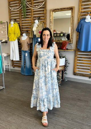 Woman modeling a sleeveless floral maxi dress with ruffle details in a boutique setting.