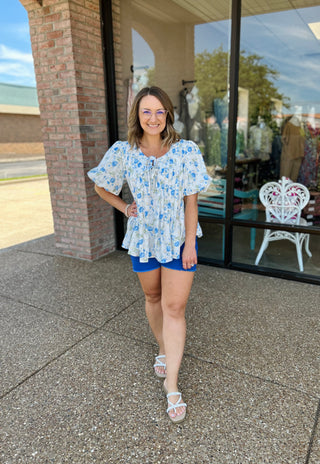 Woman wearing blue floral bubble sleeve top with smocked detail, standing outside storefront.
