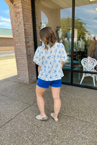 Woman wearing white floral bubble sleeve top with blue shorts, standing outside storefront