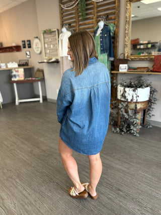 Woman wearing oversized denim shirt dress, shown from behind in boutique setting with rustic decor.