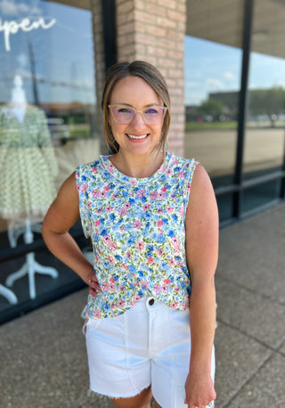 Woman wearing floral sleeveless tank top with white shorts, standing outdoors and smiling at camera.