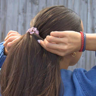 Woman styling hair with patterned scrunchie hair tie, wearing colorful beaded bracelets and blue shirt.