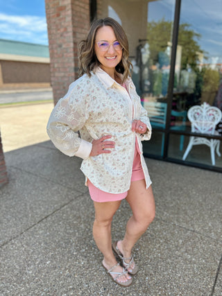 Woman wearing floral patchwork button-down shirt with pink shorts, standing outside storefront smiling.
