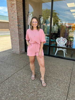 Woman wearing pink short-sleeve top and matching drawstring shorts standing outside storefront