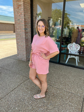 Woman wearing pink matching short set outfit standing outside storefront with brick columns