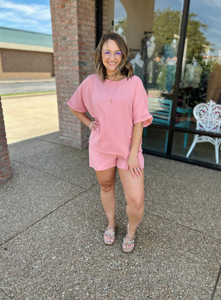 Woman wearing pink matching short set outfit standing outside storefront with glasses and sandals