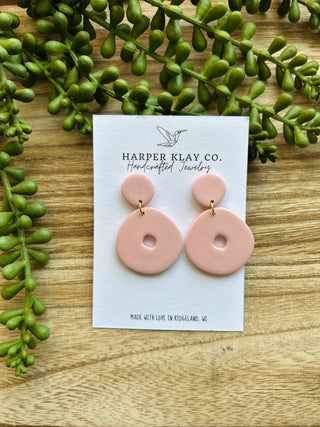 Pink clay circle earrings on branded card surrounded by green foliage on wood surface
