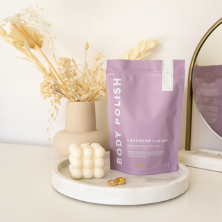 Bonblissity Lavender Luxury Body Polish moisturizing exfoliant pouch displayed on marble tray with decorative candle and d...