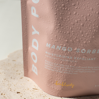 Bonblissity Mango Sorbet Body Polish Scrub pouch with water droplets on sandy surface.