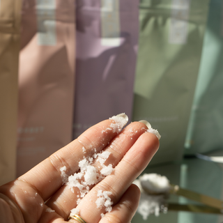 Hand holding white granular body scrub with skincare bottles blurred in background