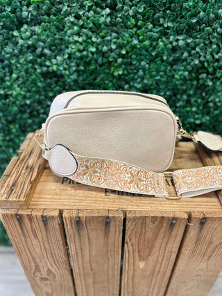 Beige crossbody bag with decorative guitar strap displayed on wooden surface with greenery background