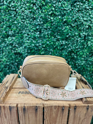 Tan crossbody bag with decorative patterned guitar strap on wooden crate against green foliage background